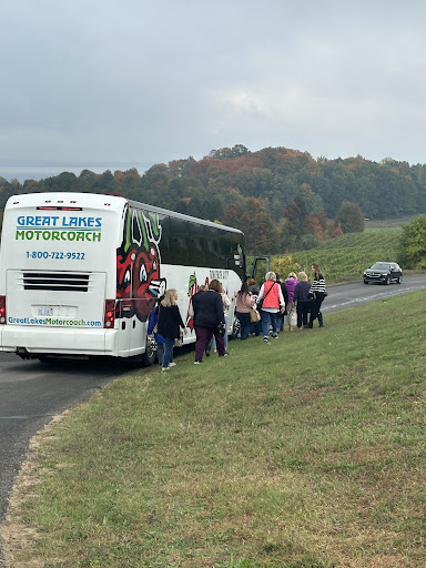 Group of passengers boarding a Great Lakes Motor Coach 56-passenger bus on a scenic road, showcasing the bus's branding and surrounding autumn landscape.